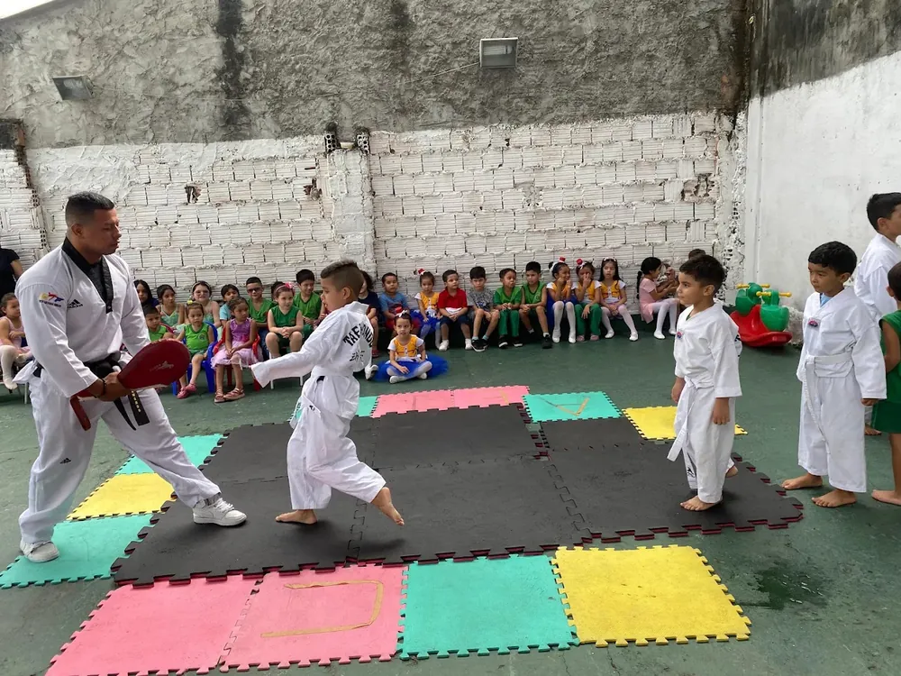 Criança aplicando um chute no escudo segurado pelo professor de taekwondo, enquanto os colegas assistem sentados.