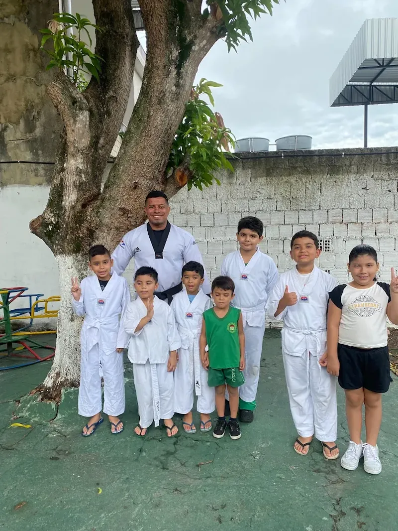 Turma de taekwondo reunida com o professor sob uma árvore, no pátio da escola.
